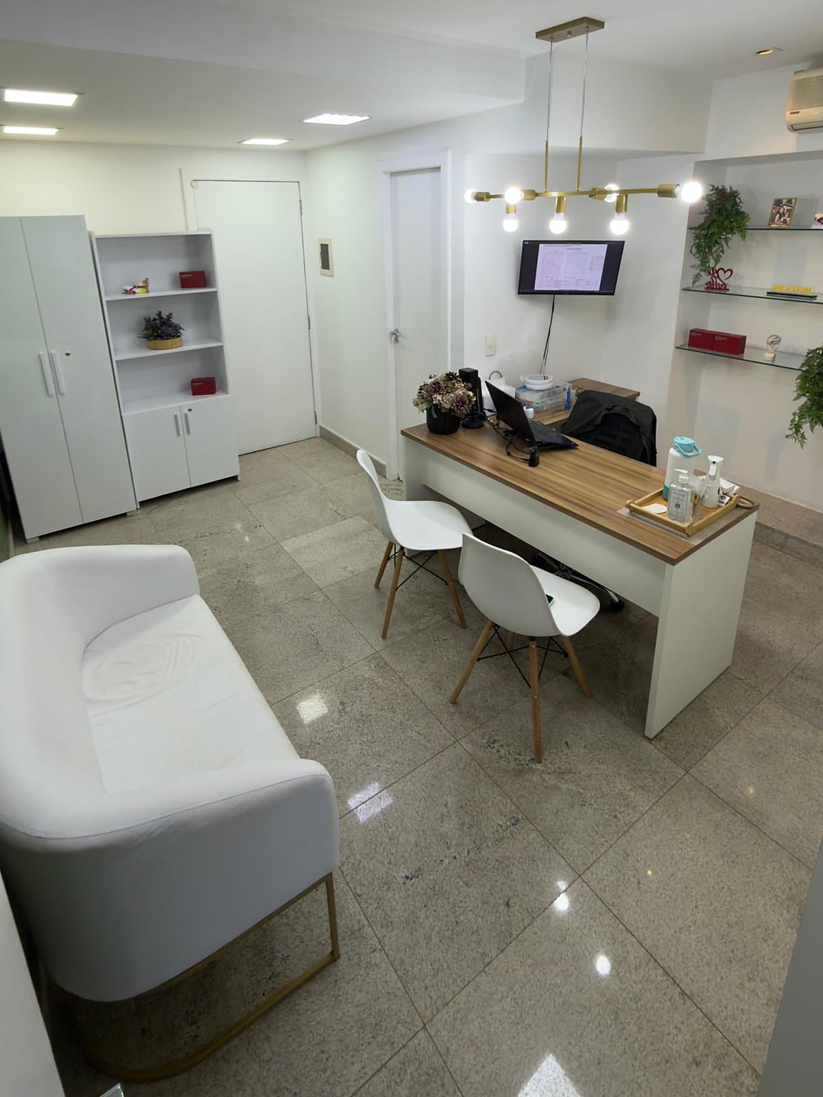 Consultório com mesa de atendimento, cadeiras, poltrona branca e monitor