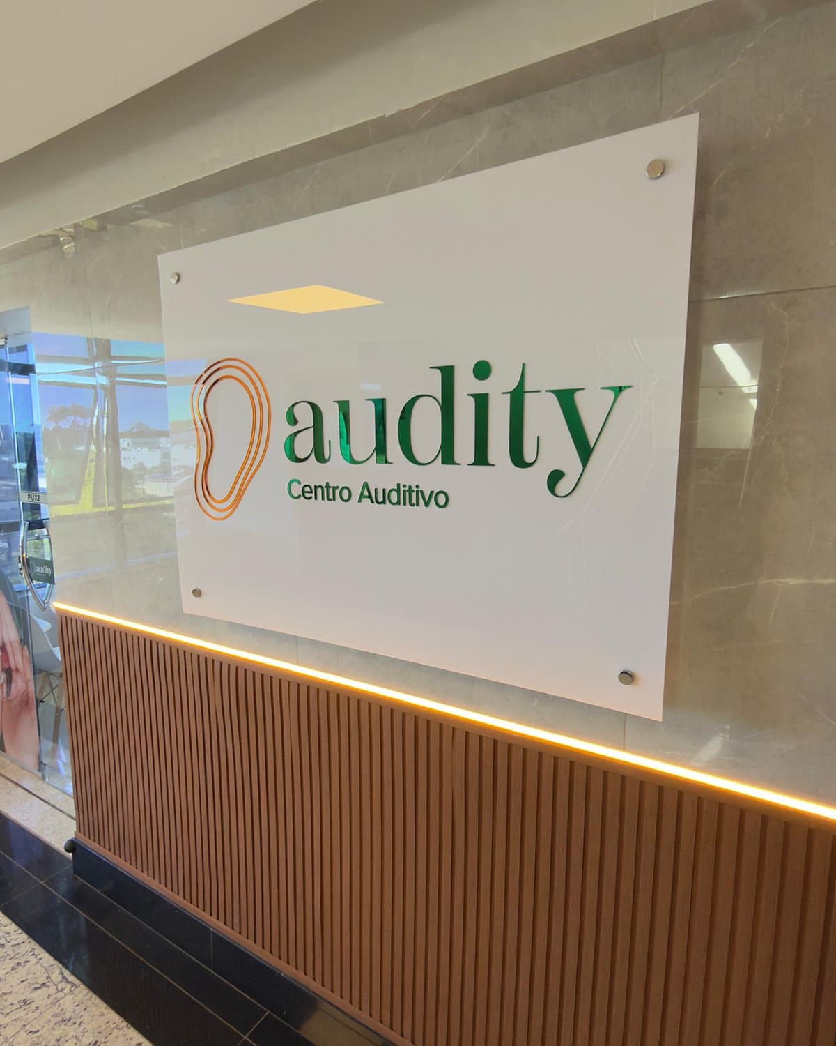 Placa da Audity Centro Auditivo no corredor da clínica, sobre painel de madeira iluminado